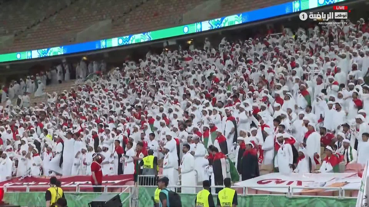 A large crowd of fans dressed in traditional white Emirati attire fills the stadium stands, cheering passionately, with "LIVE," "Abu Dhabi Sports 1," and "Abu Dhabi Sports TV" visible in the top right corner broadcast overlay, capturing the vibrant atmosphere of the Khaleeji27 final between UAE and Saudi Arabia.