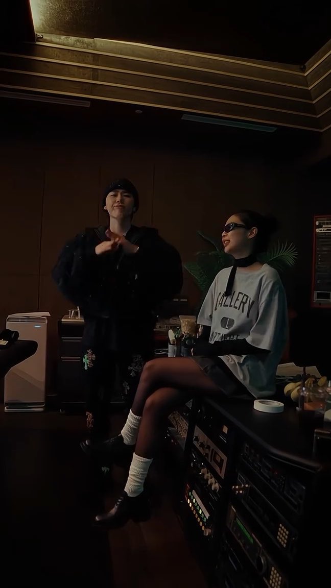 Two people pose in a dimly lit recording studio; a young man stands making a hand gesture, while a young woman in a white 'GALLERY DEPT' shirt and sunglasses sits on equipment racks.