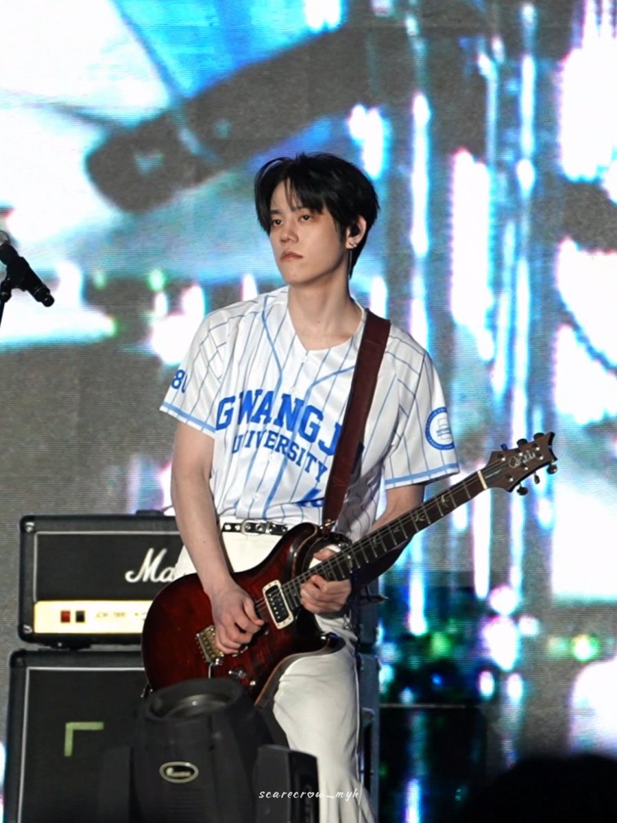 A young man with dark hair plays a red electric guitar on stage, wearing a white and blue pinstriped 'GWANGJU UNIVERSITY' jersey, with a Marshall amplifier behind him and a watermark reading 'scarecrow_myk'.