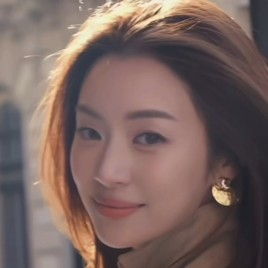 A close-up portrait of a young woman with shoulder-length brown hair featuring blonde highlights, wearing gold earrings and a tan jacket, smiling softly.