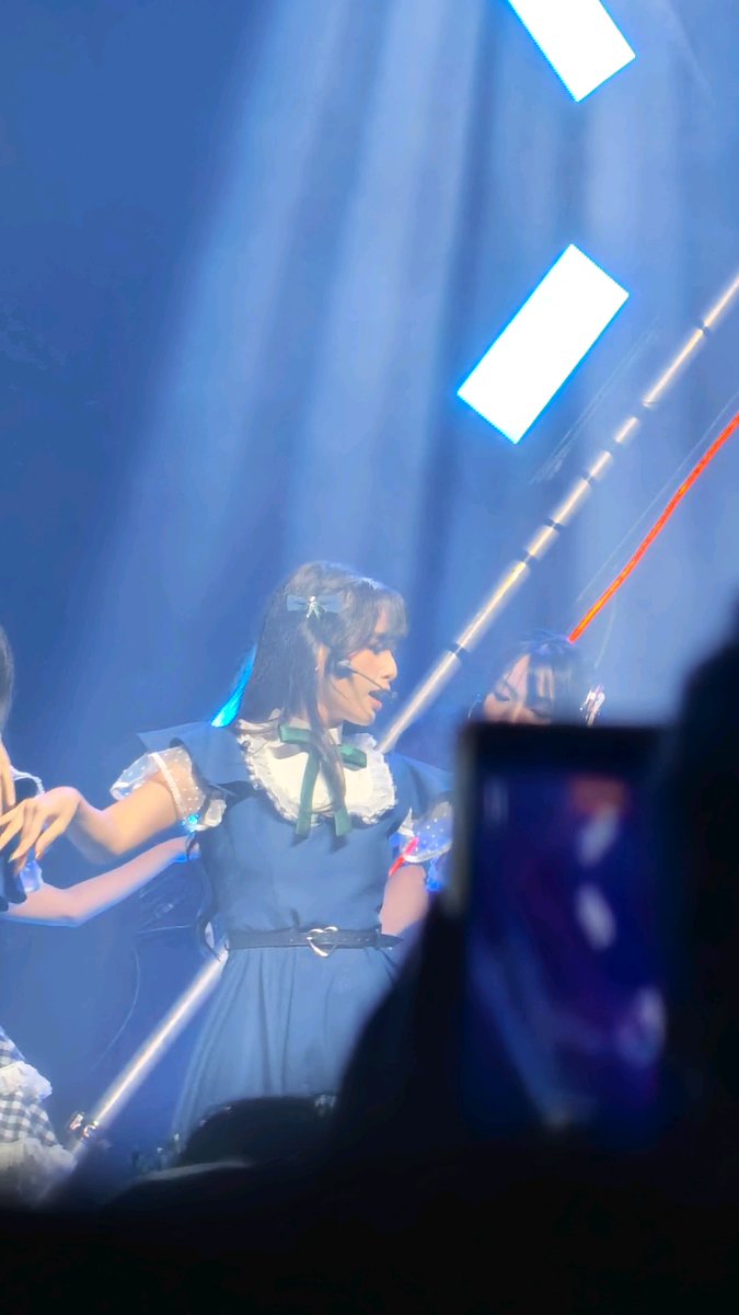 A photograph taken from an audience member's perspective at a concert, showing a performer with dark hair in a blue dress with white lace details singing on stage under blue lighting, with a phone screen visible in the foreground.