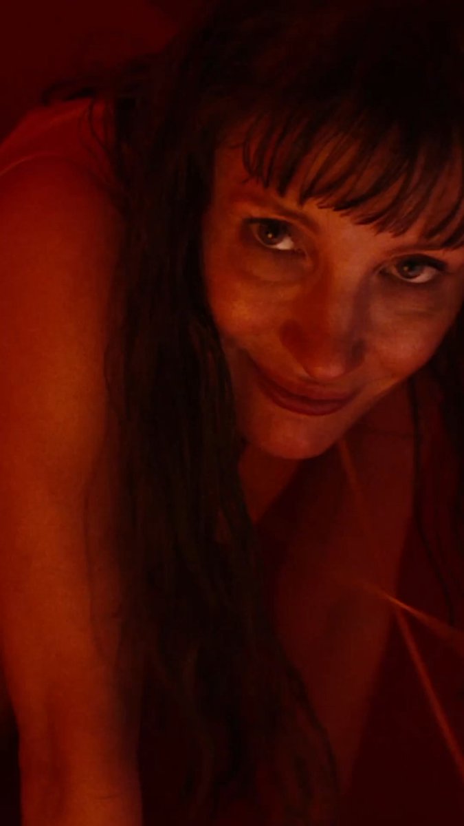 A close-up portrait of a woman with long dark hair and bangs, bathed in warm red lighting.