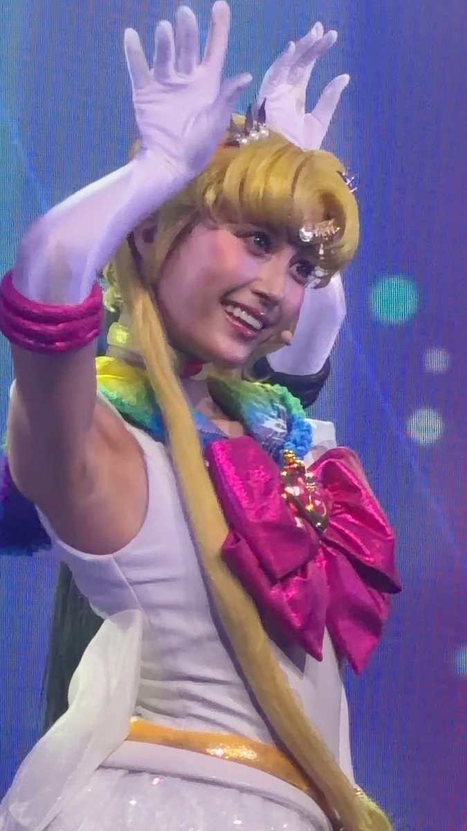 A performer in a Sailor Moon costume posing with both arms raised, wearing long white gloves and a pink bow.