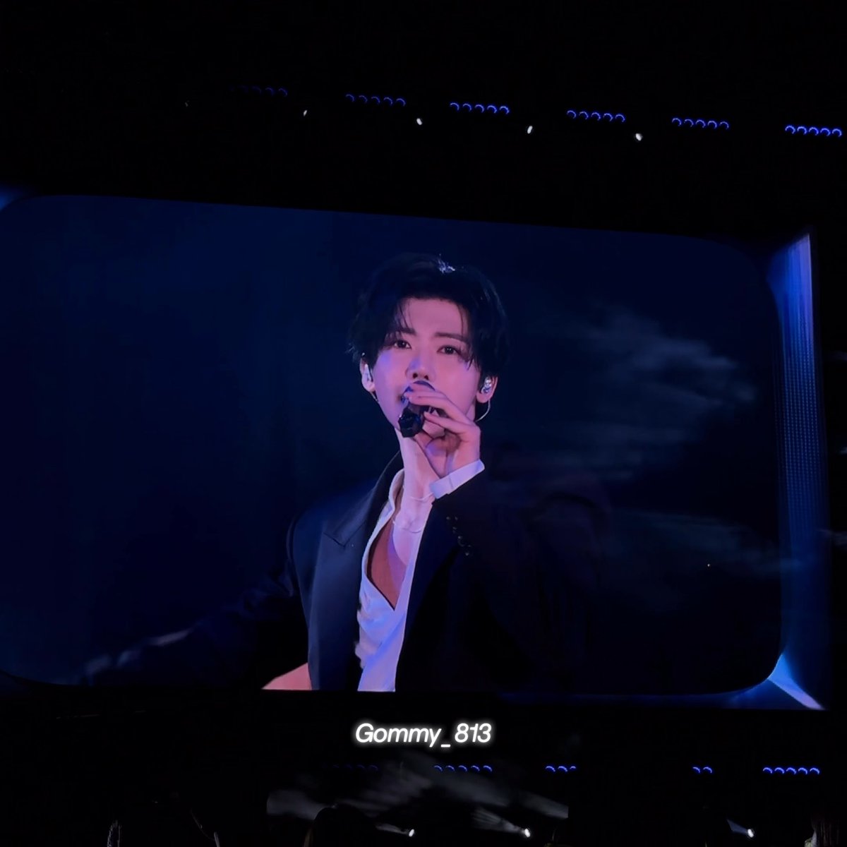 A photo taken at a concert showing a performer on a large screen singing into a microphone, wearing a suit with an open white shirt, with the username "Gommy_813" overlaid.