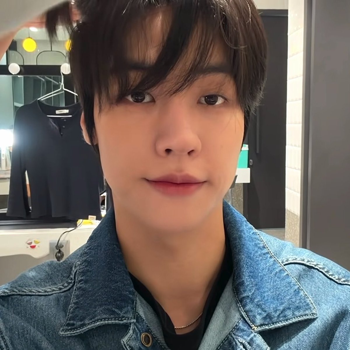 A close-up selfie shows a young man with dark hair and a denim jacket looking directly at the camera, with a black shirt hanging on a rack in the background.