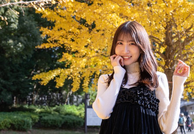 A smiling young woman wearing a black dress over a white turtleneck sweater, standing in front of bright yellow autumn trees.