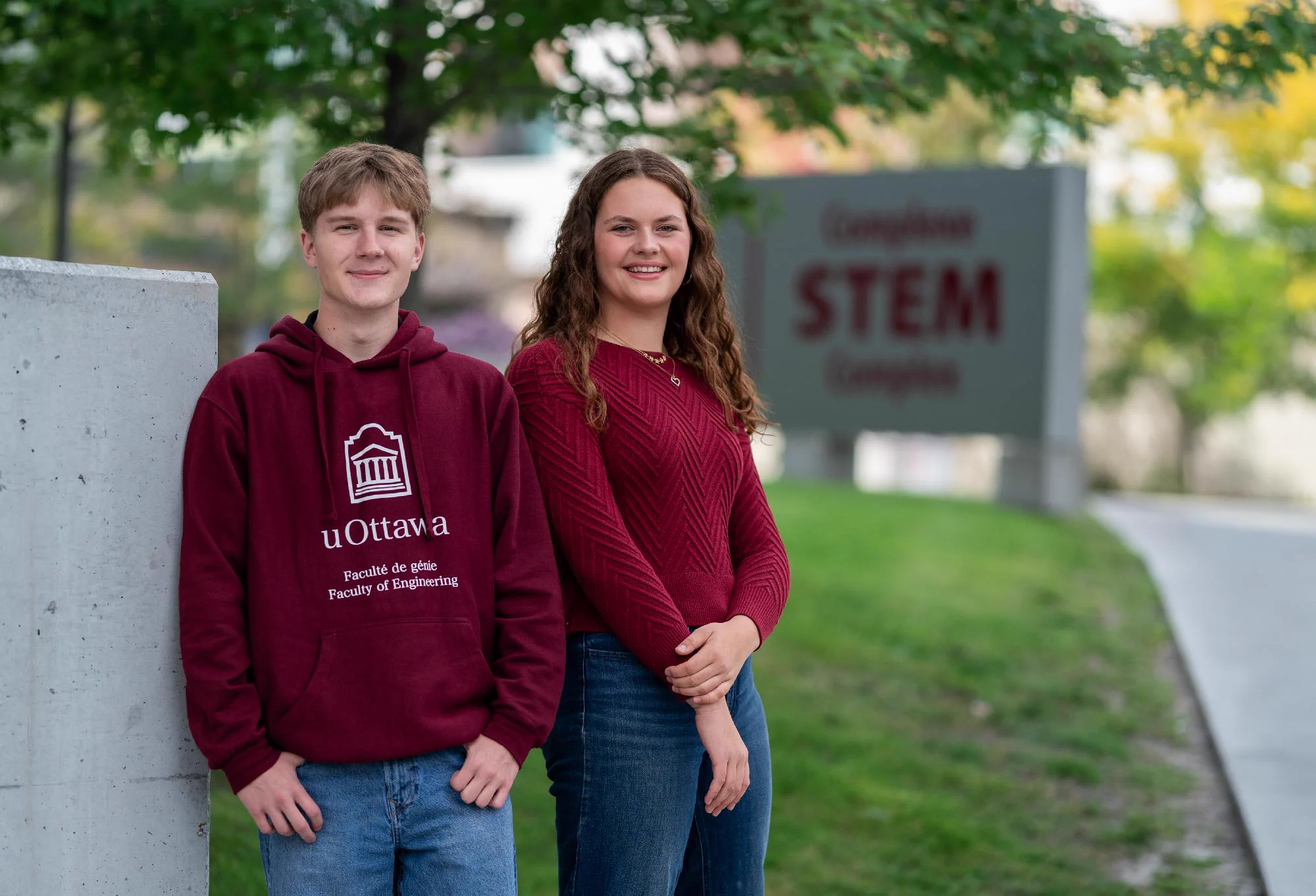 William Lane (left), student in electrical engineering and Alexandria McCully (right), student in chemistry.. They are standing outdoors near a concrete sign. William is wearing a uOttawa Faculty of Engineering sweatshirt. Alexandria is wearing a red shirt. The sign in the background reads "Complex STEM Complex." The scene is set with greenery and a pathway.