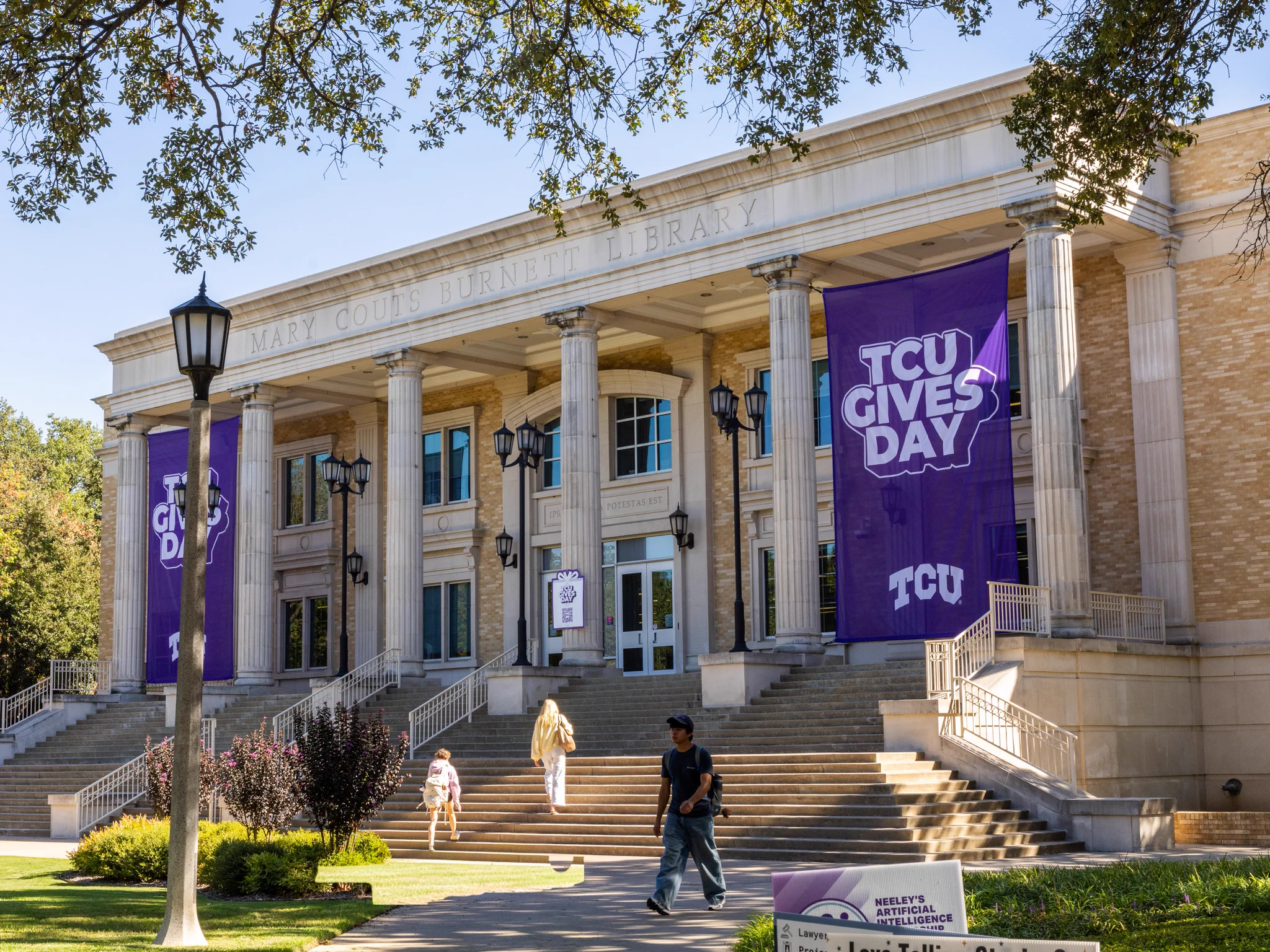 Exterior of Mary Couts Burnett Library with two large "TCU Gives Day" banners hanging from columns. Several people are walking on the steps. A lamppost and surrounding trees are visible in the foreground.
