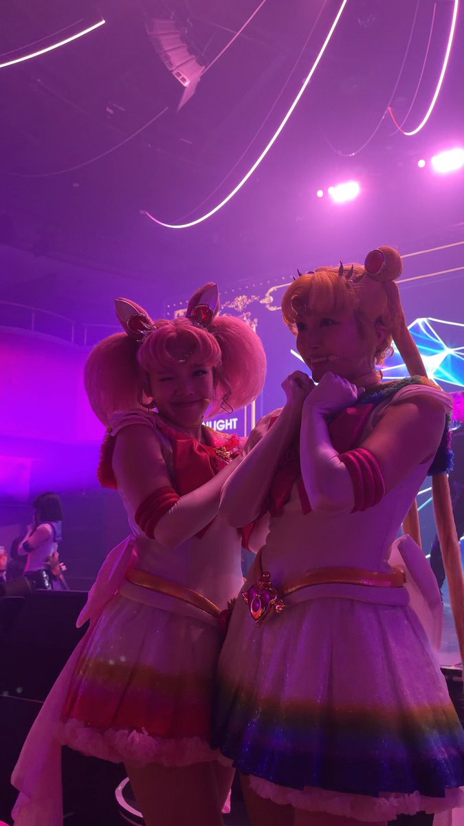 Two cosplayers dressed as Sailor Moon and Chibi Moon pose cheek-to-cheek, playfully miming blowing kisses to the camera under vibrant purple and pink lighting at the Shining Theater (STST) event in Shinagawa. The hashtag "#STST#シャイニングシアター" and date 20260910 visible in the post align with the energetic live-performance atmosphere captured in the photo.