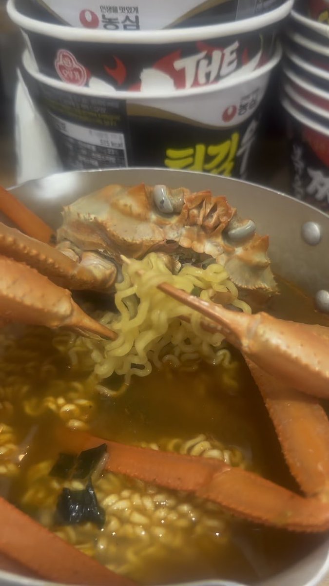A close-up of a Korean ramen cup noodle bowl containing crab, crab legs, and noodles in broth, with Korean text visible on the packaging.