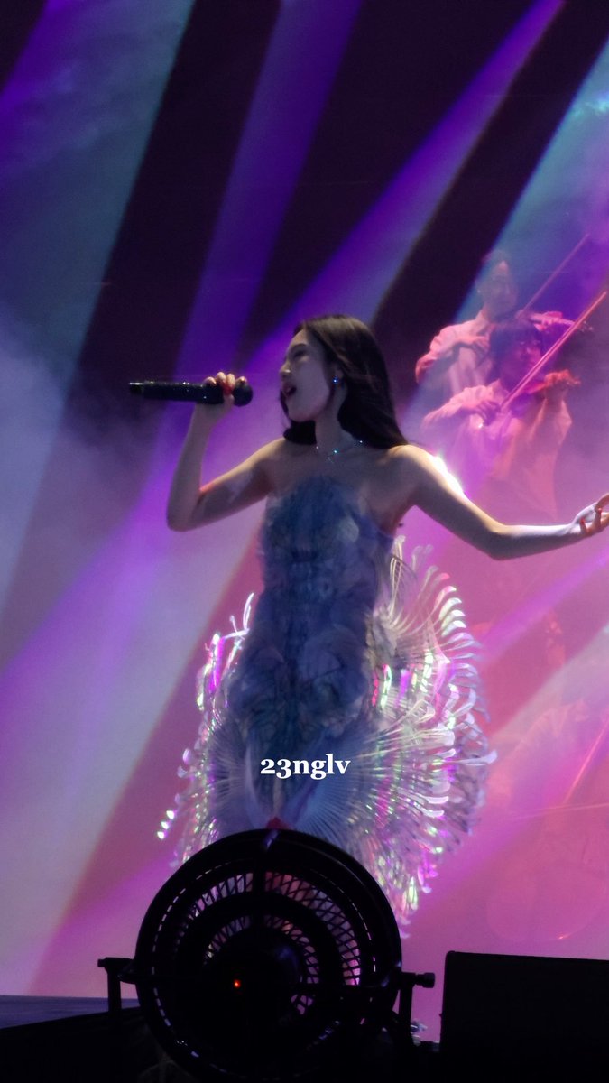 A female performer in an elaborate blue feathery dress singing on stage under purple lighting, with a violinist visible in the background and '23nglv' watermark.