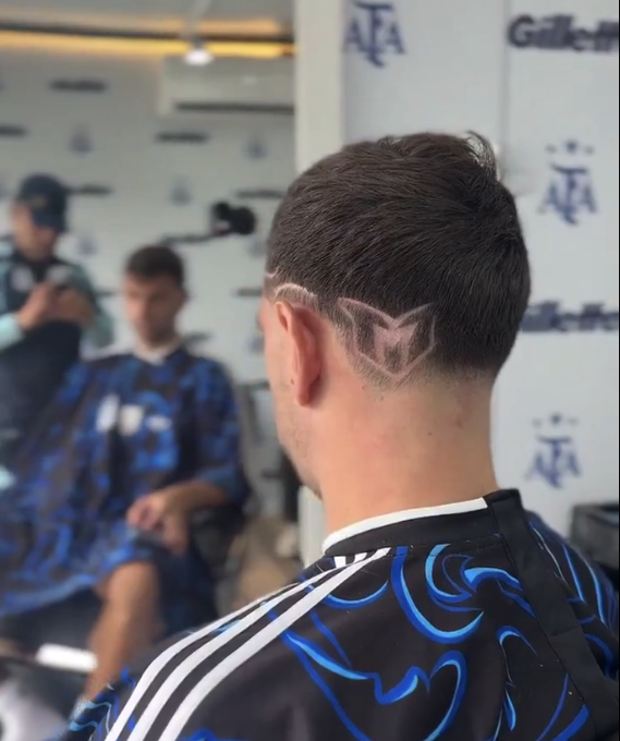 The image shows the back of a man's head featuring a stylish haircut with a shaved letter "M" design at the nape, viewed in what appears to be a barbershop with mirror reflections of other patrons. In the background, signage displays "Gillette" and "AFA" branding, along with the letters "MT" visible on the subject's blue and black patterned barber cape—though the post identifies the man as Emiliano Martínez paying tribute to Lionel Messi with the "M" shaved design.