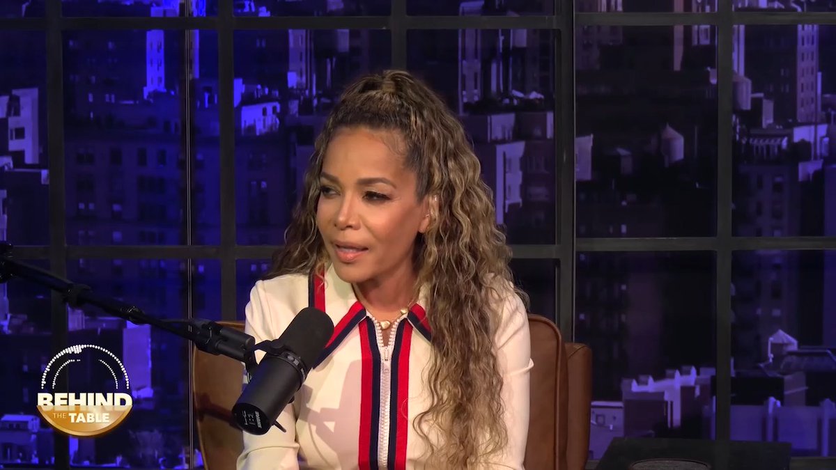 A woman with long curly hair, wearing a white blouse with red and navy stripes, speaks into a microphone during a podcast recording. The "Behind the Table" logo is visible in the bottom left corner, set against a backdrop of a nighttime cityscape seen through large windows, suggesting this is from a recent episode discussing the Cornell 7 controversy.