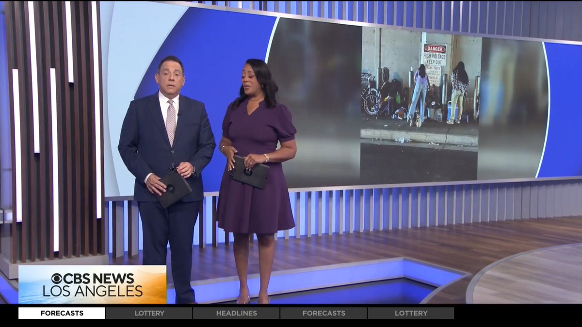 Two news anchors stand in a CBS News Los Angeles studio, with a screen behind them showing an image labeled 'DANGER HIGH VOLTAGE KEEP OUT' near a sidewalk scene, and ticker labels including FORECASTS, LOTTERY, and HEADLINES visible at the bottom.
