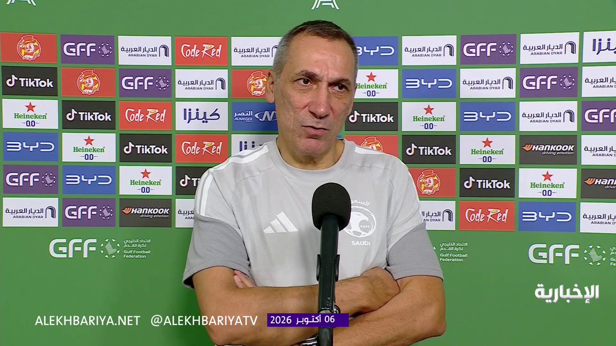 Georgios Donis (appearing in a gray Saudi national team shirt) speaks at a press conference with a backdrop featuring sponsor logos including TikTok, Heineken, Code Red, and BYD, with the date "06 أكتوبر 2026" displayed on screen.