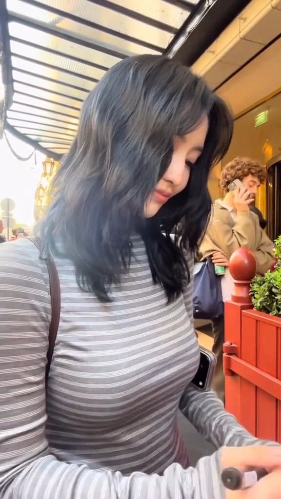 A photograph of an Asian woman with long dark hair wearing a grey striped long-sleeve shirt outdoors, with another person visible in the background talking on a phone.