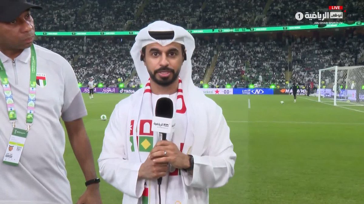 A reporter in traditional Emirati attire holds a microphone on the pitch at a football stadium, with broadcast logos for Abu Dhabi Sports and a scoreboard showing "KSA 00" visible in the background, while the caption indicates he is interviewing Ismail Rashid, the UAE national team manager, about the team's preparations ahead of the Gulf Cup 27 final against Saudi Arabia.