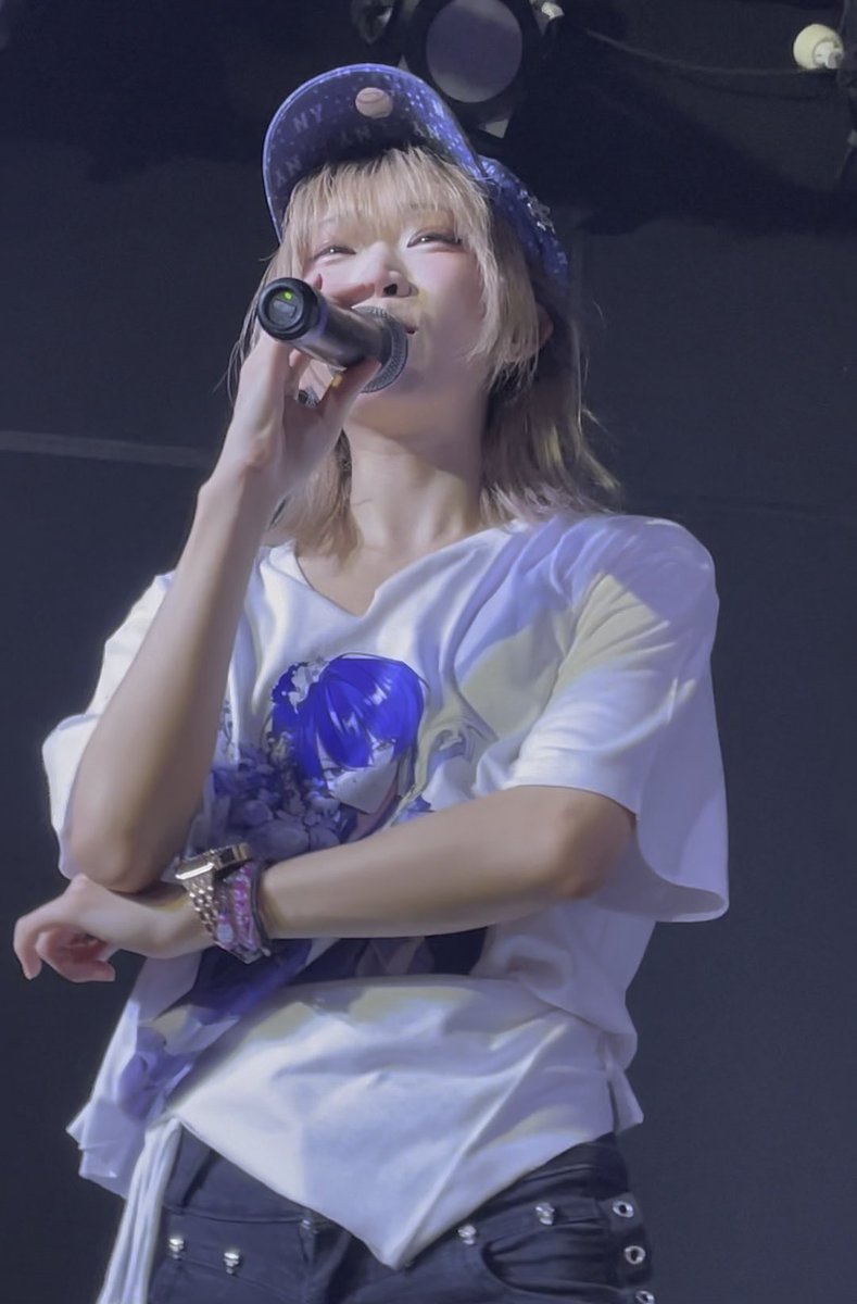 Image 2: A female performer with short blonde hair, wearing a galaxy-print cap and a white anime-themed t-shirt, sings into a microphone on stage under concert lighting.