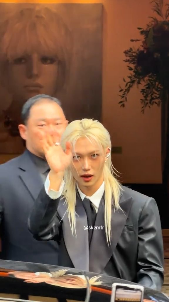 A person with long blonde hair in a dark suit waving their hand, with an older man behind them and a painting in the background; a watermark reads '@skzmfr'.