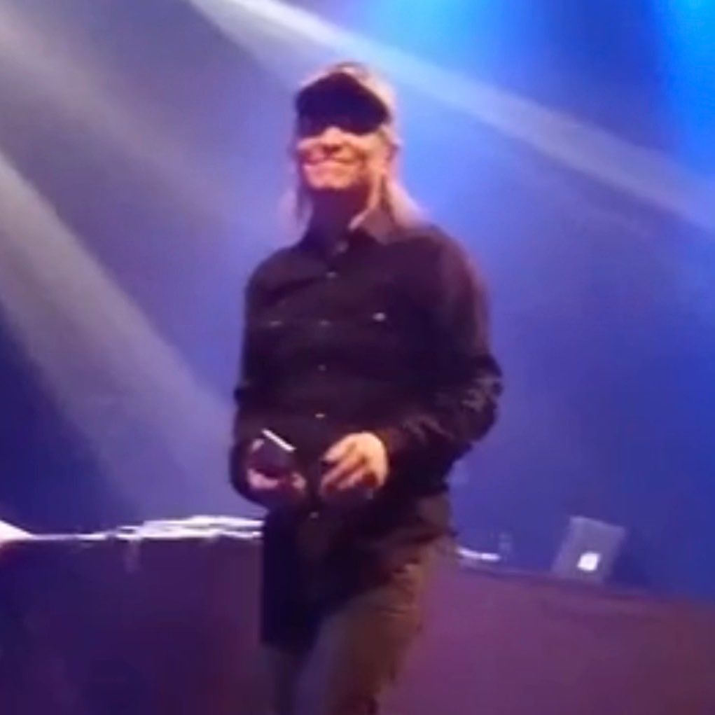 A blonde man wearing sunglasses and a black jacket stands on a stage illuminated by blue and white diagonal lights.