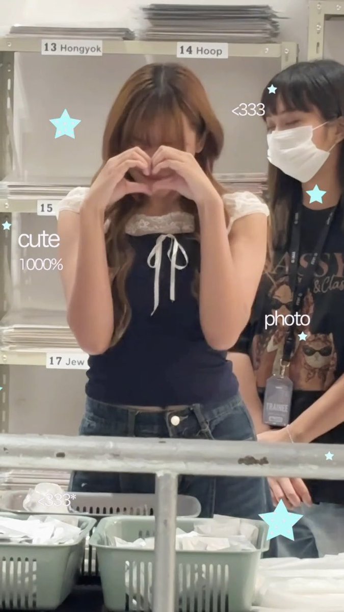 A young woman with brown hair forms a heart shape with her hands in front of her face, wearing a navy blue top with white lace trim, in what appears to be a workspace with numbered labels '13 Hongyok', '14 Hoop', and '15' visible in the background.