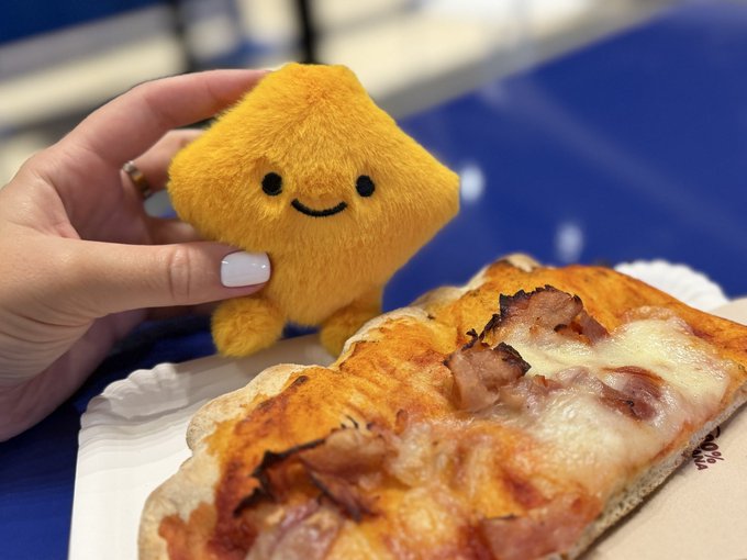 A hand holds a small, smiling yellow star plushie next to a slice of cheesy flatbread pizza with bacon on a paper plate.