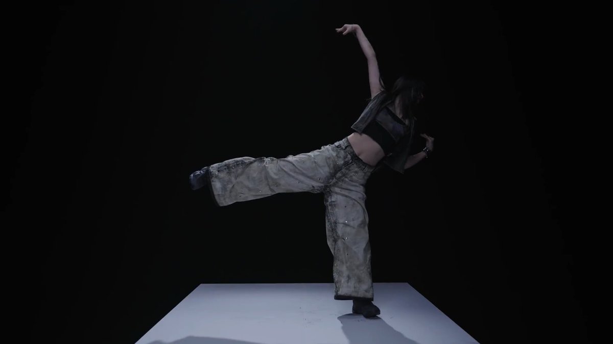 A dancer performs on a white platform against a black background, wearing loose, distressed grey pants and a black vest while executing a high leg extension.