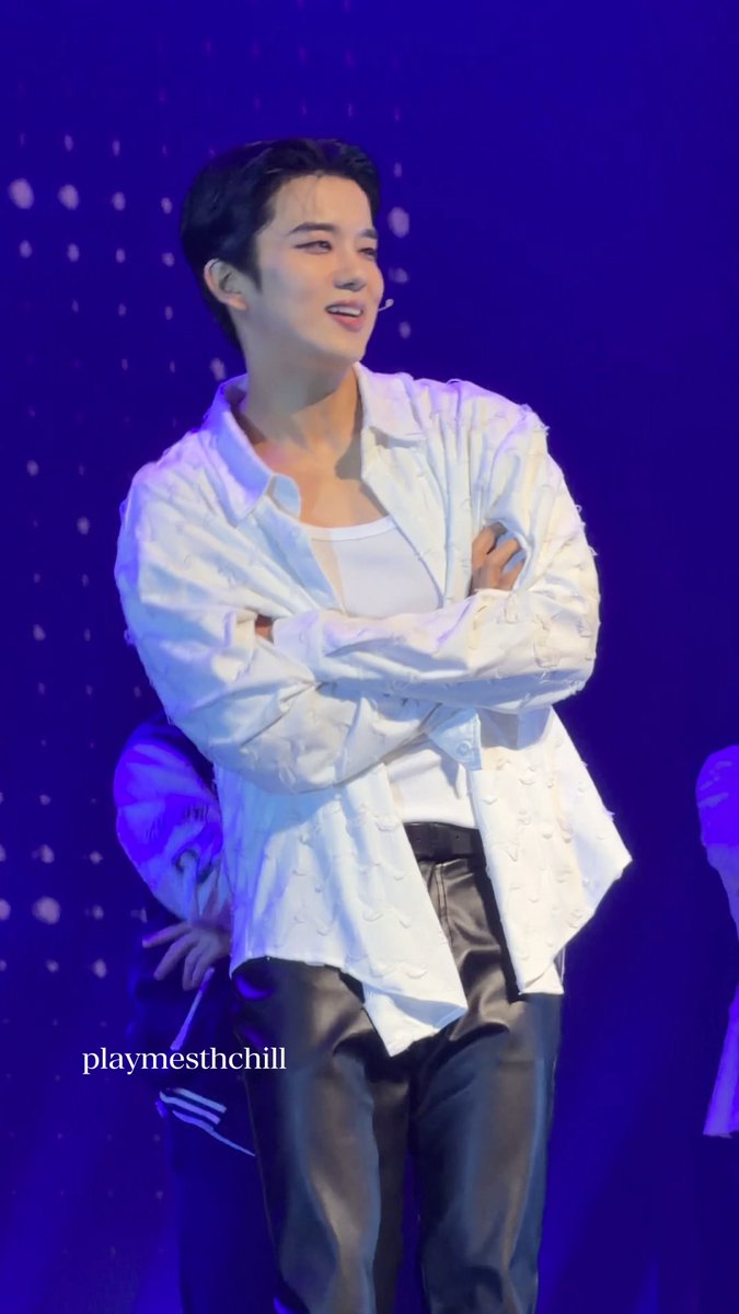 A male performer with dark hair, wearing a white patterned shirt over a white tank top and black leather pants, smiles on stage under blue lighting with the text 'playmesthchill' visible.
