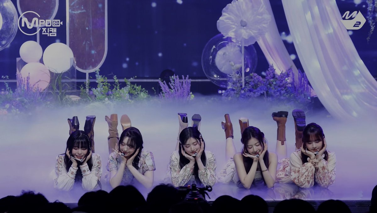 A K-pop girl group performing on stage at MPD Fan Cam, with five members lying on their stomachs in coordinated poses amid purple lighting and lavender decorations.