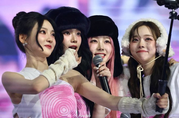 Photo of four K-pop idols, including members wearing furry hats, performing on stage with microphones, with a watermark reading 'Satellite'.