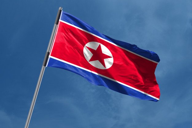 The flag of North Korea waves against a blue sky, featuring its characteristic red, white, and blue stripes with a red star in a white circle.