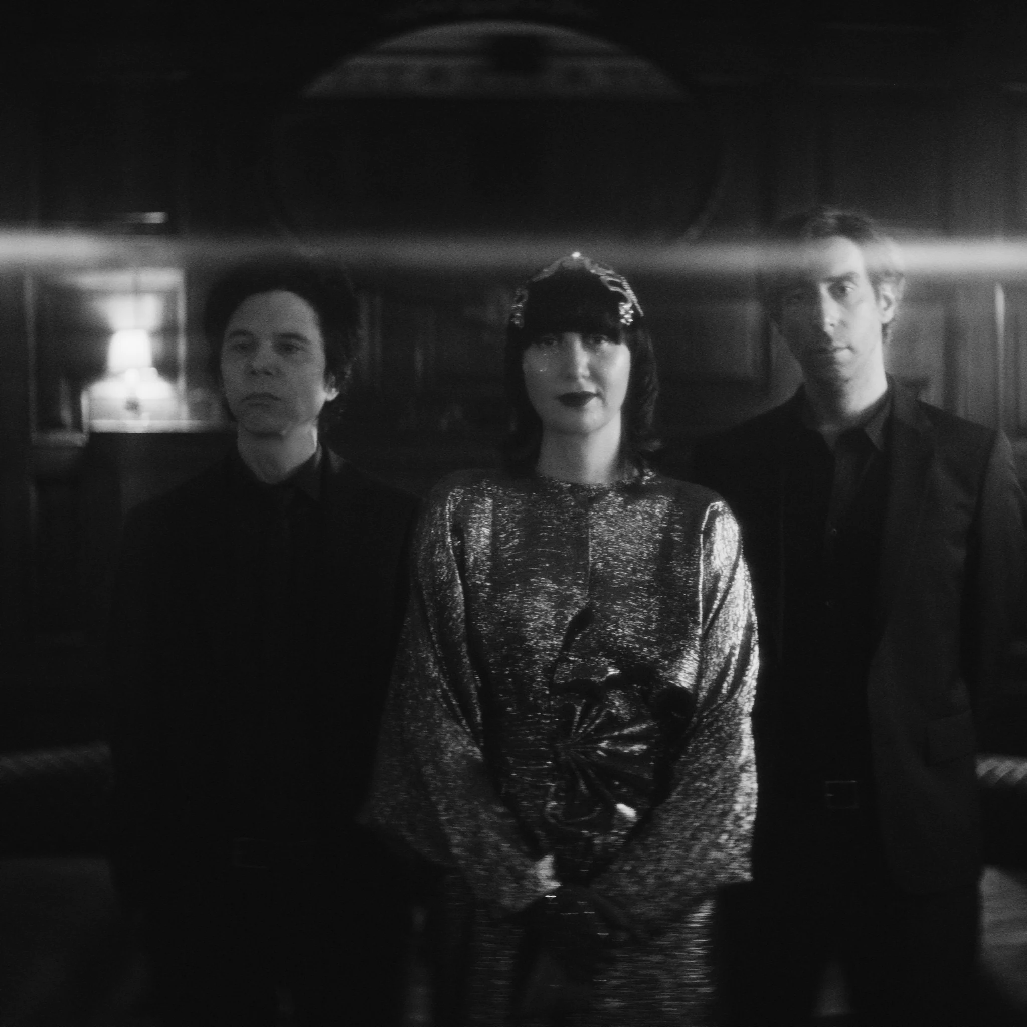 A black-and-white photo of the Yeah Yeah Yeahs standing together in a dim room, with Karen O centered in a shimmering dress and dark gloves between Nick Zinner and Brian Chase.