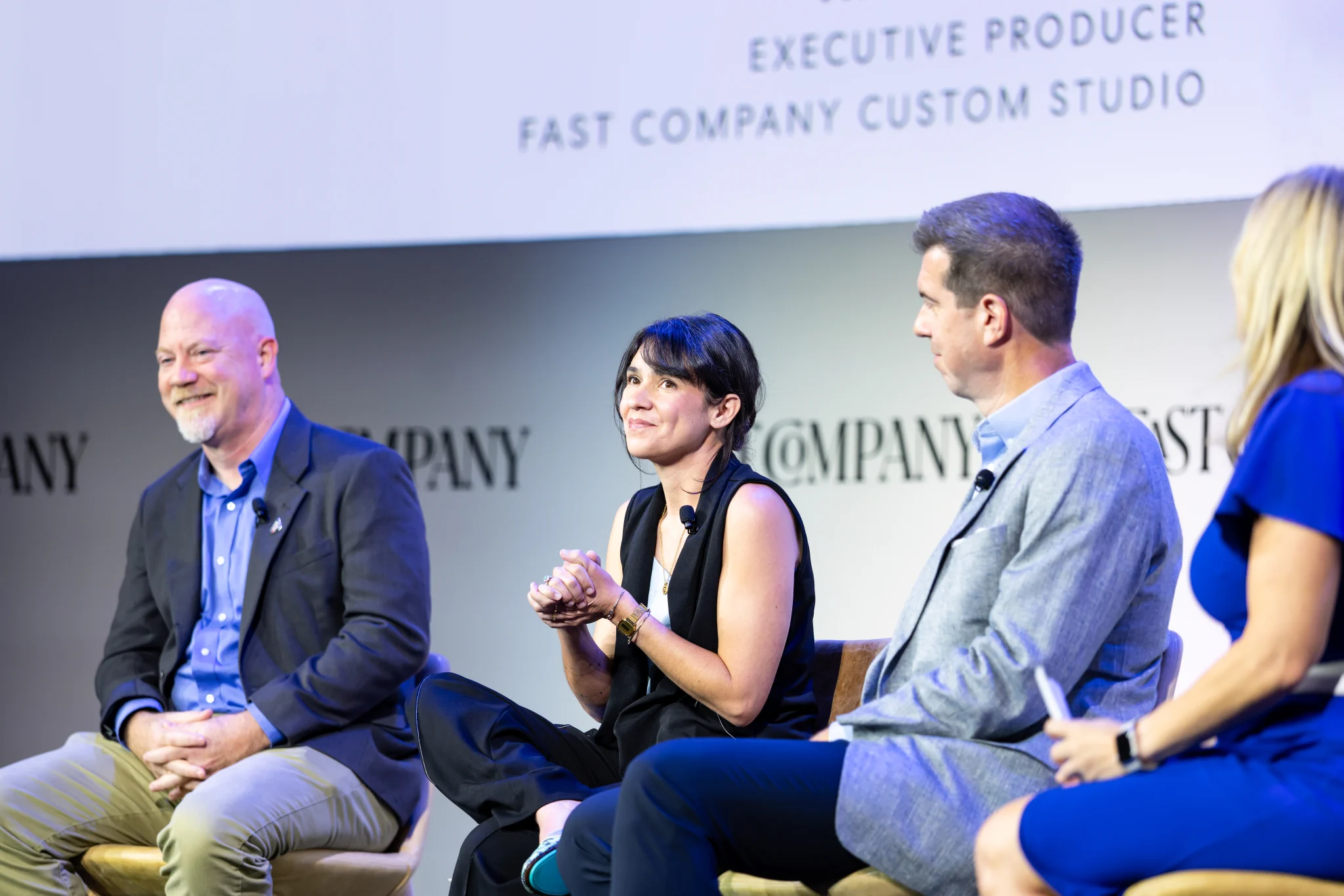 Three people are seated on a stage at a Fast Company event, engaged in discussion. A fourth person is partially visible, sitting beside them. The backdrop displays the Fast Company logo and information about an executive producer. The atmosphere is professional and conversational.