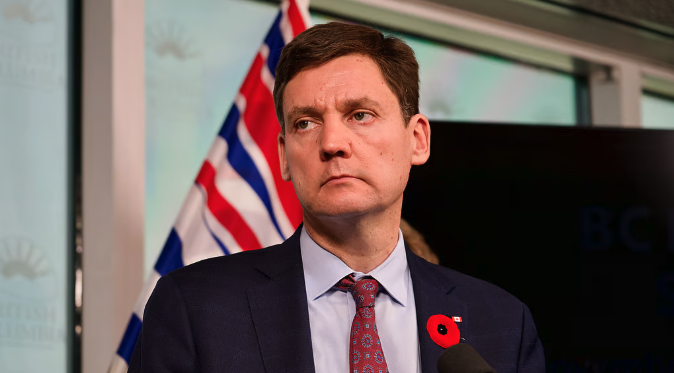 A stern-looking man in a dark suit and patterned tie wearing a red poppy pin stands before a British Columbia flag and what appears to be a BC government backdrop, with partial text "BC" visible. The image relates to the post mocking David Eby, the British Columbia Premier, suggesting his anticipated majority victory amid Conservative Party turmoil has not materialized as hoped.