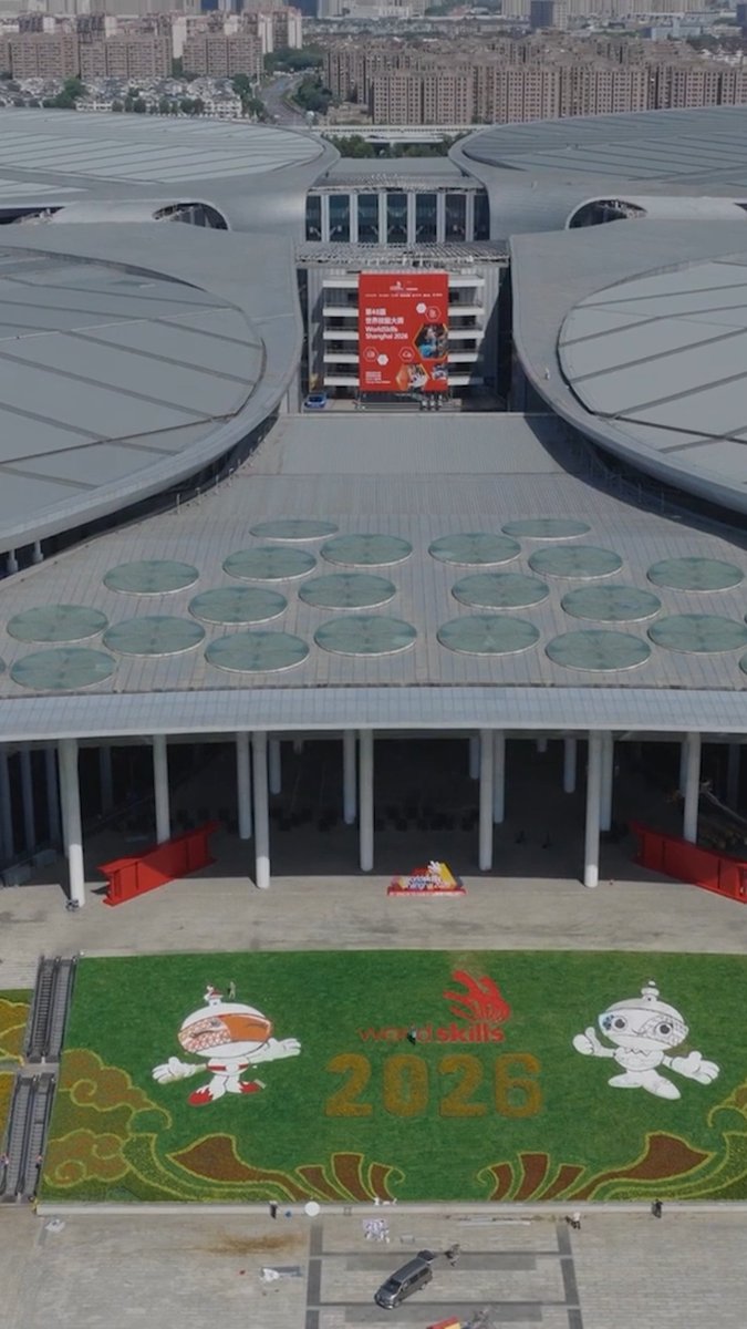 An aerial view of the Chongqing International Expo Center during WorldSkills 2026, featuring a large lawn display with the event's mascot characters and "world skills 2026" text, along with a red banner reading "WorldSkills Chongqing 2026" hanging between the venue's distinctive circular roofed pavilions. The image showcases the impressive venue where the CNC Milling and Turning competitions have begun, setting the stage for the skilled competitors shaping manufacturing's future.