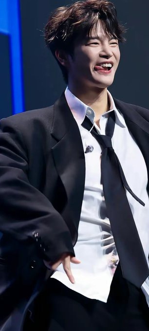 A young man with dark hair laughing while wearing a black blazer over a white shirt with a loose black tie, likely a K-pop idol at an event.