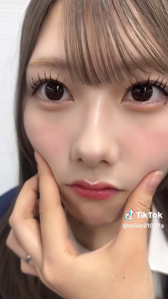 Close-up TikTok selfie of a young woman (@eriierii1027a) with dramatic doll-like eye makeup, long lashes, pink blush and glossy lips, framing her face with both hands.