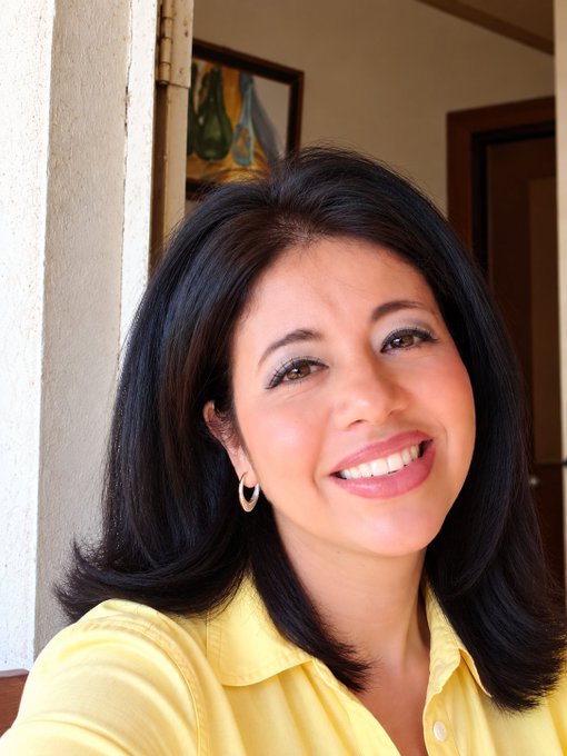 A portrait photo of a smiling woman with dark hair wearing a yellow polo shirt and hoop earrings.