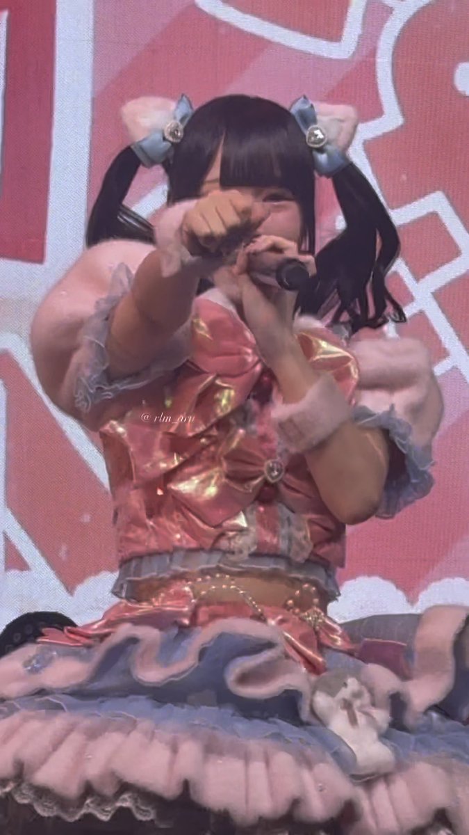 A live concert photo of a Japanese idol performer in an elaborate pink and lavender frilly idol costume with twin pigtails, singing into a microphone while wiping her eyes on stage; the image is credited to '@rin_air' (@ri_ain).