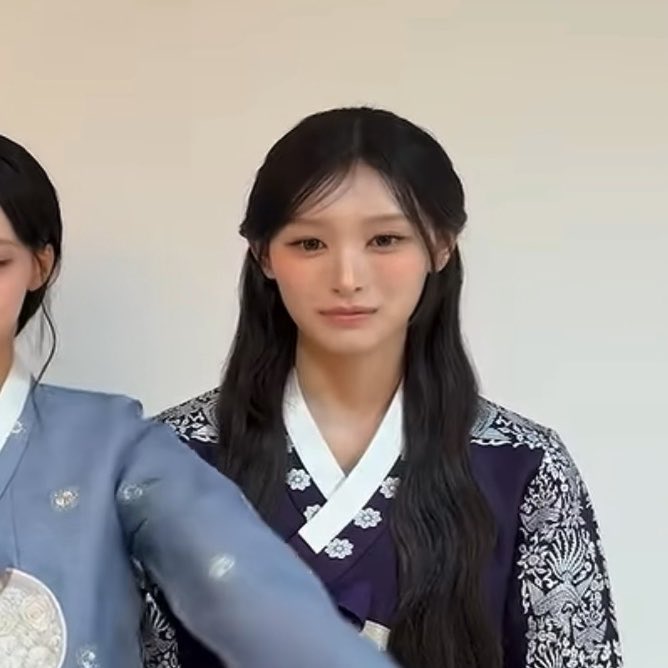 A young woman with long dark hair wearing a purple and white traditional Korean hanbok smiles at the camera, with another person partially visible to the left.