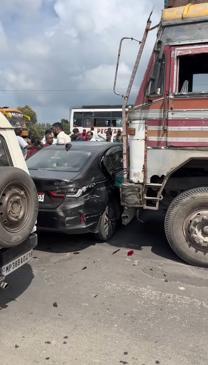 A photo of a traffic accident showing a damaged black sedan sandwiched between a large decorated Indian truck and another vehicle, with onlookers and a bus in the background.