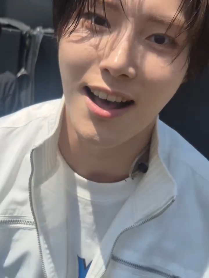 A close-up selfie of a young man with messy dark hair wearing a white zip-up jacket over a white t-shirt, his mouth slightly open as he looks into the camera in bright sunlight.