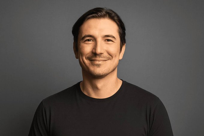A portrait photograph of a smiling middle-aged man with short dark hair, wearing a black t-shirt against a plain gray background.