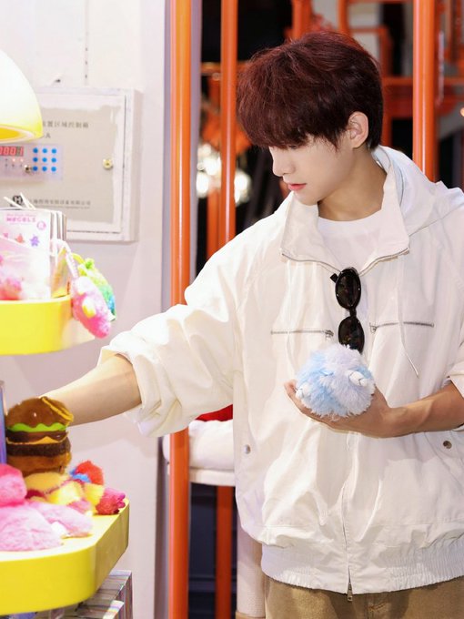 A young man with reddish-brown hair, wearing a white jacket and sunglasses hanging from his shirt, looks at a colorful display of plush toys.