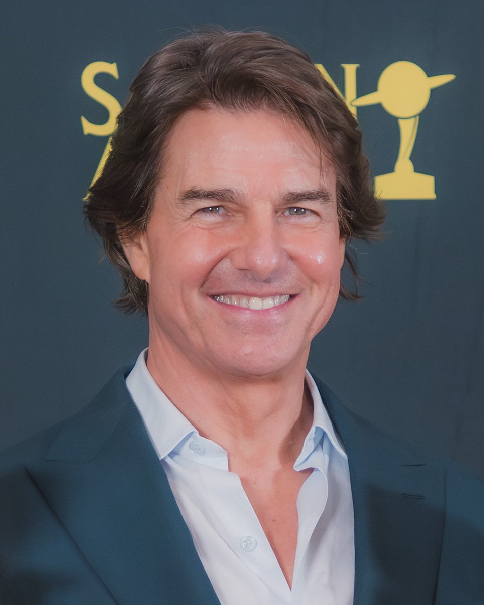 Tom Cruise smiling at an awards event, wearing a white shirt and dark blazer, with a gold Saturn Award logo behind him.