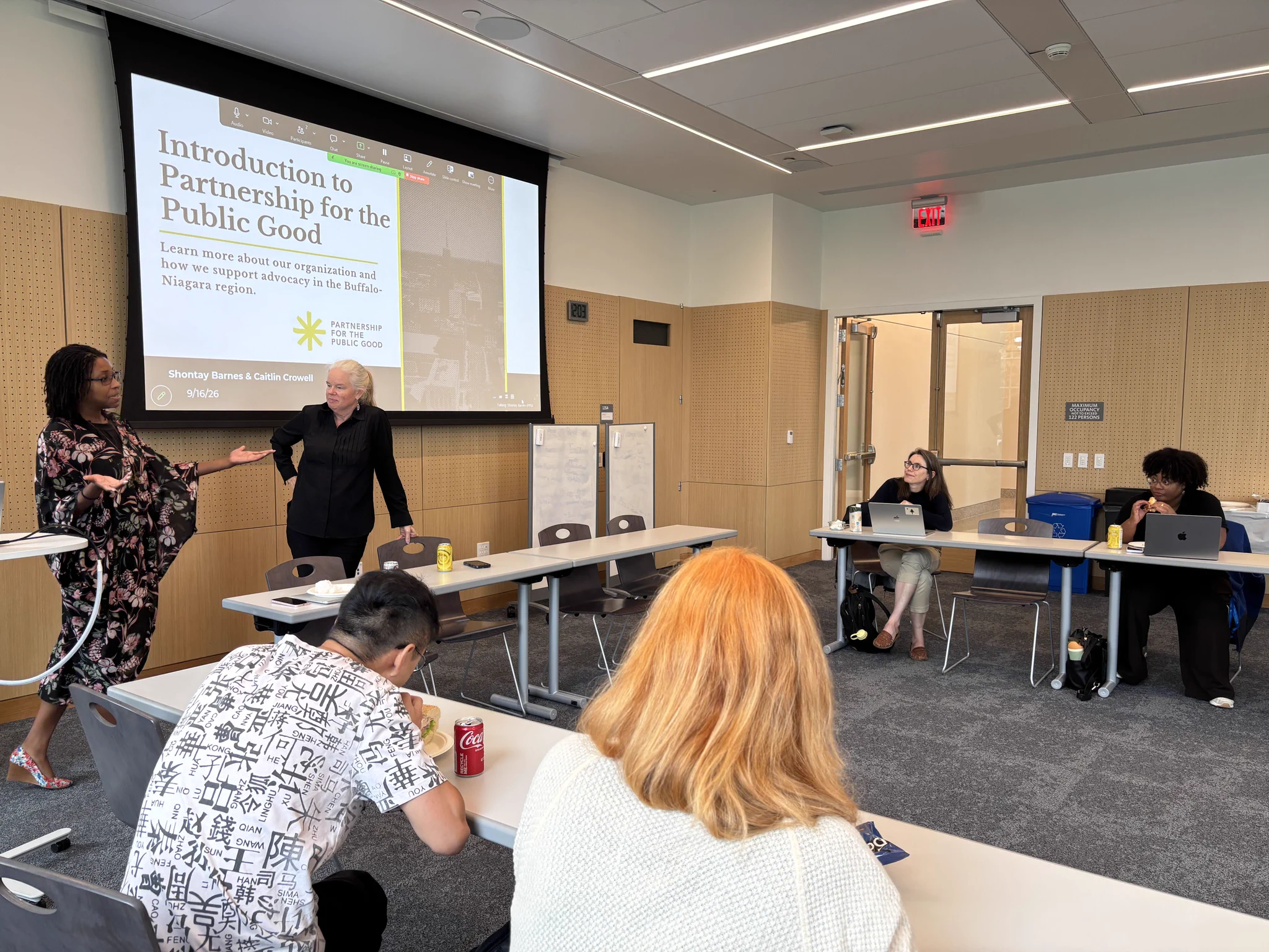 Brightly lit classroom: two women, Shontay Barnes & Caitlin Crowell, present \"Introduction to Partnership for the Public Good.\" One speaks gesturing, the other listens. Several attendees sit at tables with laptops, drinks (Coca-Cola, yellow can), and snacks, some eating. Clock reads 12:03.