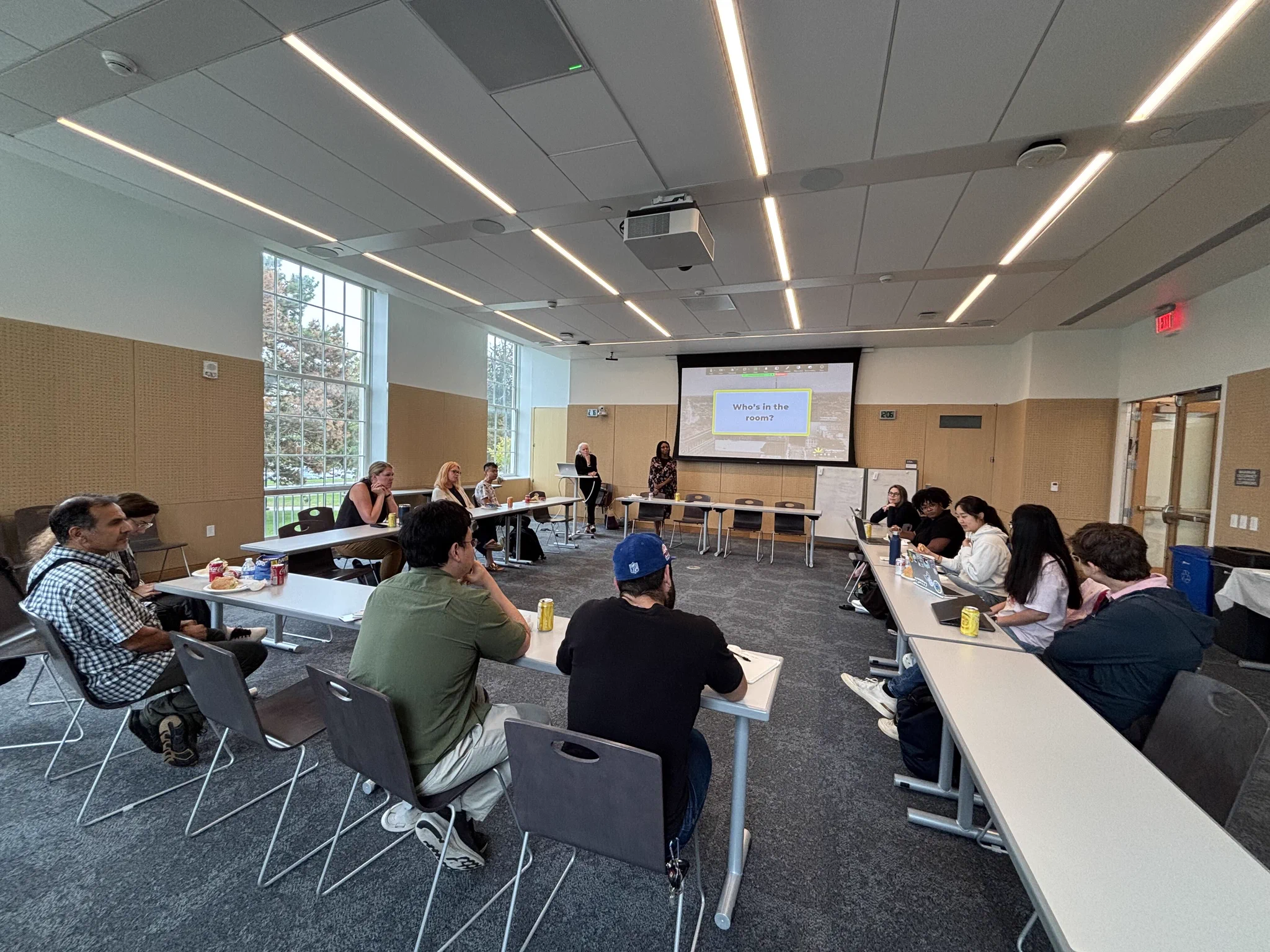 A diverse group of adults attends a meeting in a bright room. Two women stand at the front near a projector screen displaying \"Who's in the room?\". Attendees are seated at long tables arranged in a U-shape, some with laptops, drinks, and snacks.