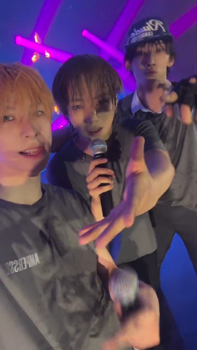Three young men pose for a selfie at a concert venue, with one holding a microphone and wearing a Yankees cap, while neon pink stage lights glow in the background.