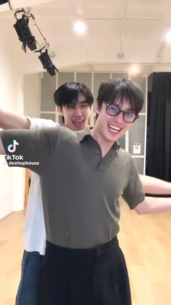 Two young men in a dance studio, one wearing glasses and a gray polo shirt, are smiling and laughing playfully for a TikTok video, with the watermark '@deehuphouse' visible.