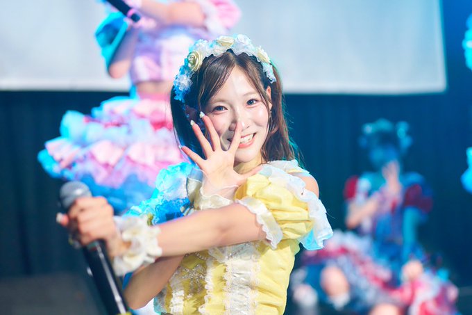A young female performer smiles brightly while singing into a microphone, wearing a yellow frilly idol costume with a decorative headpiece, with other performers visible in the background.